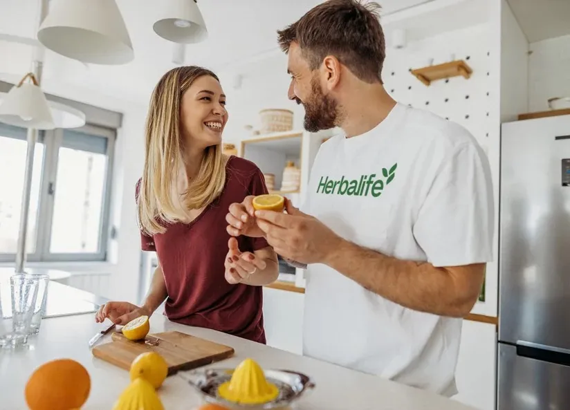 Pareja sonriendo en la cocina preparando cítricos, con camiseta Herbalife
